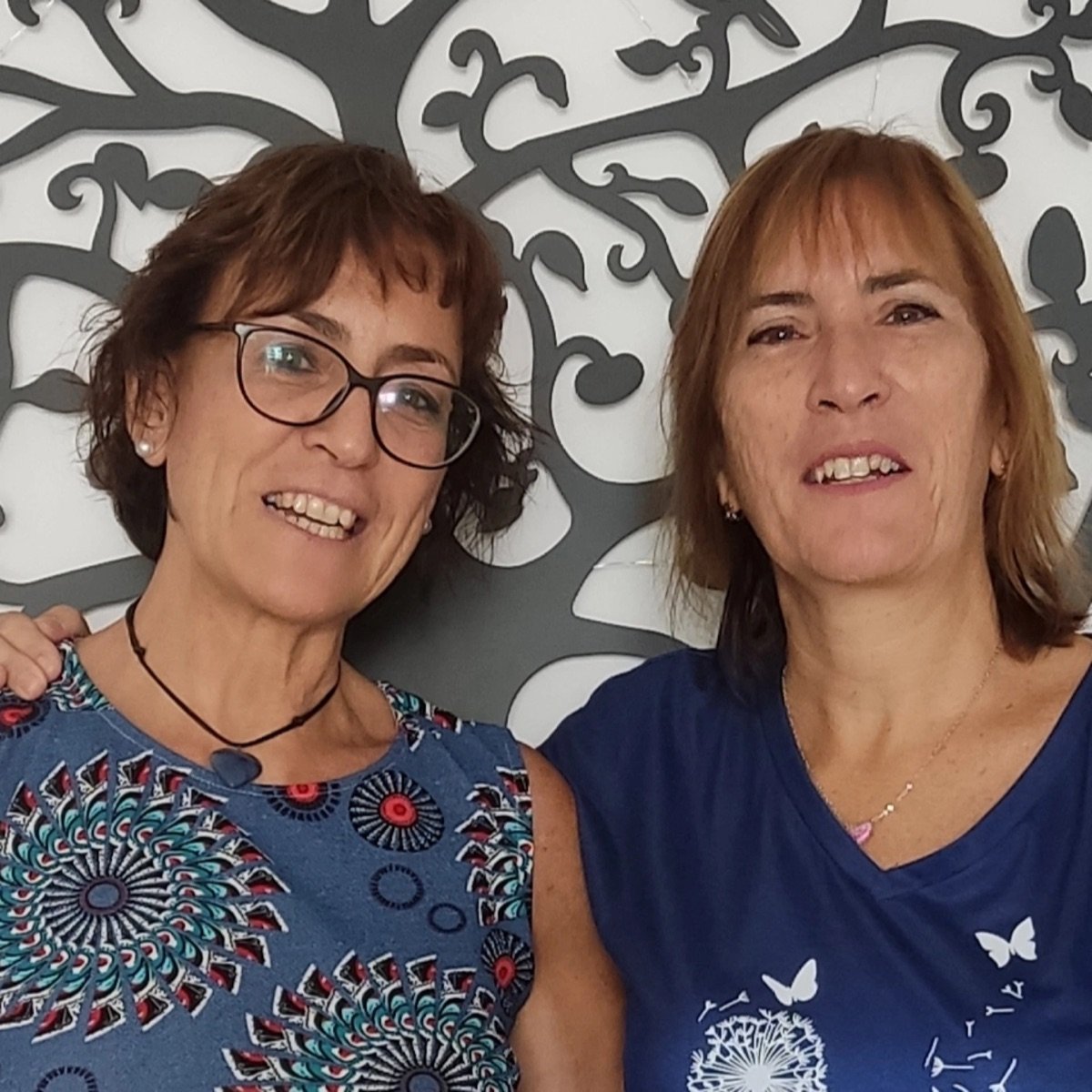 Retrato cercano de Maribel y Sonia, sonriendo, frente a un árbol de la vida.